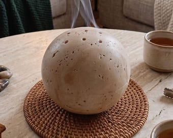Può includere: Un bruciatore di incenso sferico color beige con una superficie porosa e testurizzata, dotato di piccoli fori sulla parte superiore. Sottili fili di fumo bianco si levano dal centro della sfera, appoggiata su un tappetino rotondo in rattan marrone.