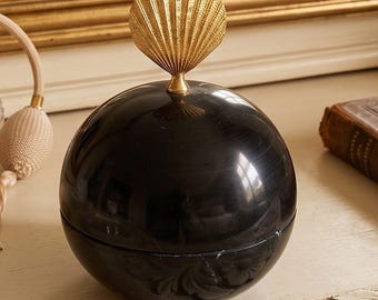 Può includere: Un contenitore sferico nero con una finitura liscia e lucida e una cucitura orizzontale. Il coperchio è sormontato da una maniglia in metallo dorato a forma di conchiglia a ventaglio con creste verticali, centrata sulla parte superiore.