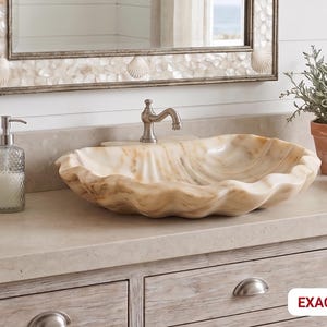 Könnte beinhalten: Ein Badezimmerwaschtisch mit einem muschelförmigen Waschbecken, einem dekorativen Spiegel und einer Topfpflanze. Das Waschbecken ist hellbeige mit gewelltem Rand. Ein Glas-Seifenspender und ein Wasserhahn sind ebenfalls sichtbar.