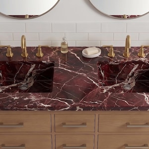 May include: A double vanity unit featuring a deep red marble countertop with white veining and two integrated rectangular sinks. The base is made of light-toned wood with six drawers and silver-toned handles. Two gold-toned taps are installed.