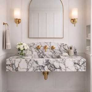 Puede incluir: Un lujoso baño con una encimera de mármol y accesorios dorados. El tocador cuenta con un lavabo integrado, un grifo dorado y un espejo arqueado a juego. Las paredes son blancas y el suelo tiene un patrón de cuadros blancos y negros.