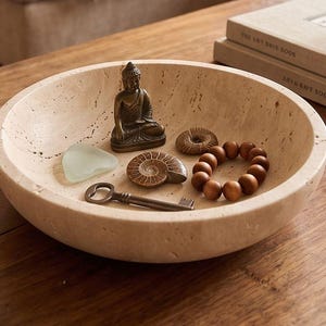 Pode incluir: Uma tigela redonda de pedra bege porosa contendo uma pequena estatueta de Buda em bronze, um pedaço de vidro marinho verde-claro, um fóssil de amonite castanho, um pequeno objeto circular texturizado, uma chave de metal e uma pulseira de contas de madeira.