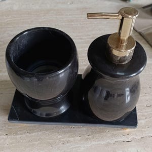 Pu&ograve; includere: Un set da bagno in pietra nera composto da un bicchiere cilindrico e un dispenser di sapone con pompa color oro, entrambi poggiati su un vassoio rettangolare in pietra nera abbinato. Il set presenta una finitura liscia e lucida su tutto.