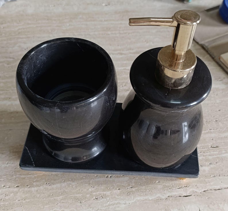Pu&ograve; includere: Un set da bagno in pietra nera composto da un bicchiere cilindrico e un dispenser di sapone con pompa color oro, entrambi poggiati su un vassoio rettangolare in pietra nera abbinato. Il set presenta una finitura liscia e lucida su tutto.
