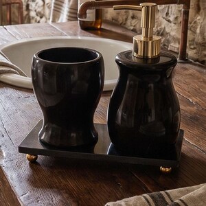 Pu&ograve; includere: Un set da bagno nero composto da un dispenser di sapone con erogatore dorato e un bicchiere abbinato, entrambi posizionati su un vassoio rettangolare nero con quattro piccoli piedini sferici dorati. Gli oggetti hanno una finitura liscia.