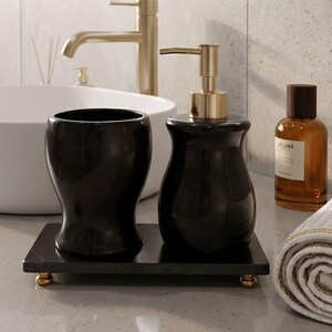 Pu&ograve; includere: Un set di accessori da bagno nero composto da un dispenser di sapone con pompa dorata e un bicchiere abbinato, entrambi poggiati su un vassoio rettangolare nero con quattro piccoli piedini dorati. Finitura liscia e lucida.