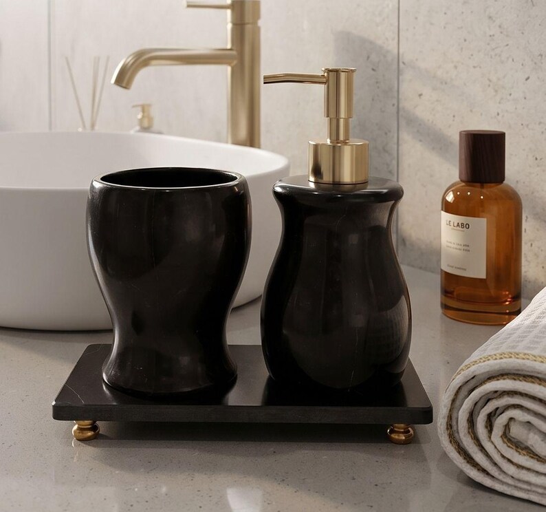 Pu&ograve; includere: Un set di accessori da bagno nero composto da un dispenser di sapone con pompa dorata e un bicchiere abbinato, entrambi poggiati su un vassoio rettangolare nero con quattro piccoli piedini dorati. Finitura liscia e lucida.