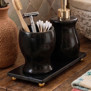 Pu&ograve; includere: Un set di accessori da bagno nero composto da un bicchiere cilindrico e un dispenser di sapone con pompa dorata, entrambi poggiati su un vassoio rettangolare nero con piccoli piedini dorati. Il bicchiere contiene uno spazzolino in bamb&ugrave;, un rasoio e cotton fioc.