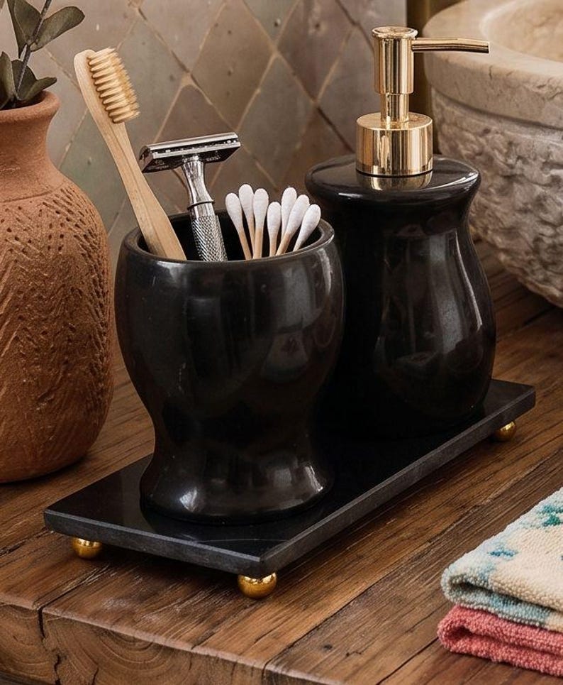 Pu&ograve; includere: Un set di accessori da bagno nero composto da un bicchiere cilindrico e un dispenser di sapone con pompa dorata, entrambi poggiati su un vassoio rettangolare nero con piccoli piedini dorati. Il bicchiere contiene uno spazzolino in bamb&ugrave;, un rasoio e cotton fioc.