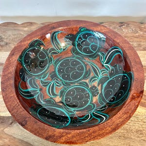 May include: A round wooden bowl with a glossy finish. The interior features a design of black sea turtles with teal and white accents. The bowl has a rich, reddish-brown color and is likely hand-crafted.