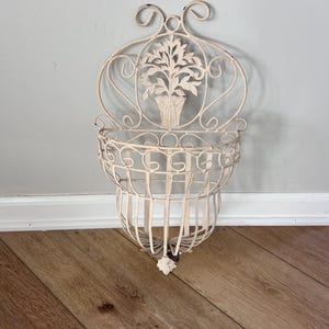 May include: Off-white metal wall planter with a decorative floral design. The planter has a basket-like structure with a decorative top and a small, ornate flower at the bottom. The wall decor is designed to hold small plants or flowers.