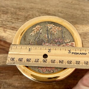 K&ouml;nnte beinhalten: Eine runde Schmuckdose aus goldfarbenem Metall mit einem dekorativen Deckel, der ein Pfauen-, Bambus- und Blumenmotiv zeigt. Ein h&ouml;lzernes Fiskars-Lineal liegt auf dem Deckel und zeigt einen Durchmesser von etwa 9 Zentimetern.