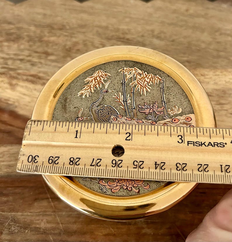 K&ouml;nnte beinhalten: Eine runde Schmuckdose aus goldfarbenem Metall mit einem dekorativen Deckel, der ein Pfauen-, Bambus- und Blumenmotiv zeigt. Ein h&ouml;lzernes Fiskars-Lineal liegt auf dem Deckel und zeigt einen Durchmesser von etwa 9 Zentimetern.