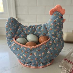 May include: A quilted fabric chicken-shaped egg warmer in a dusty blue color with a small floral pattern in pink and green. The chicken has a peach-colored felt comb and beak, with a small dark eye. It holds three eggs in a central pouch.