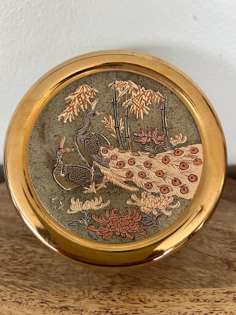 K&ouml;nnte beinhalten: Runde, goldfarbene Zierschale mit detailliertem Pfau- und Blumendesign. Die Mitte zeigt zwei Pfauen, Bambus und Blumen in Braun-, Orange- und Wei&szlig;t&ouml;nen auf gr&uuml;nem Hintergrund.
