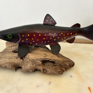 May include: A hand-painted wooden fish sculpture with a dark body, red and yellow spots, and black fins. The fish rests on a piece of driftwood, showcasing a rustic and natural aesthetic. The fish has green eyes.