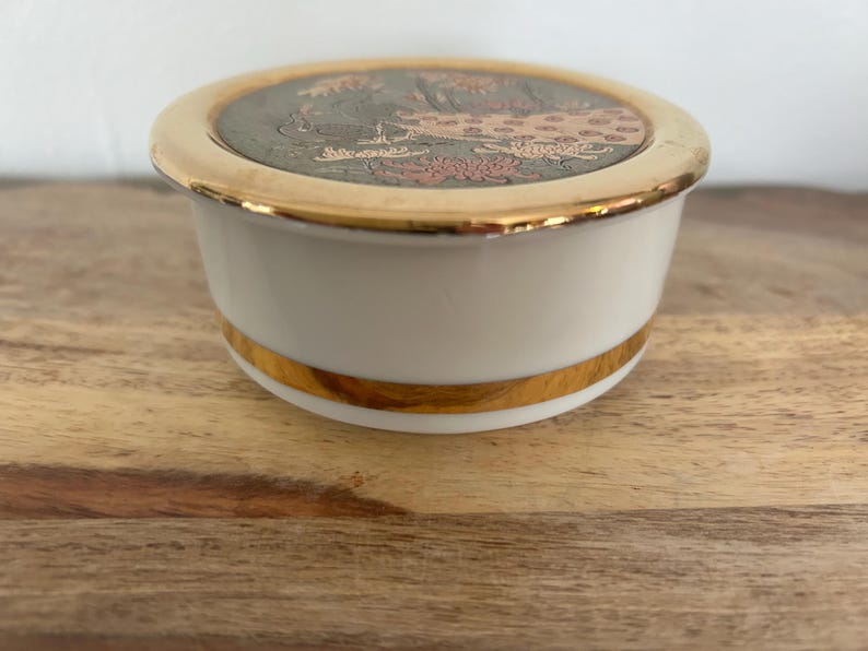 K&ouml;nnte beinhalten: Eine runde Schmuckdose aus wei&szlig;em Porzellan mit goldfarbenem Rand an Deckel und Boden. Der Deckel hat eine dunkle, kreisf&ouml;rmige Einlage mit einer detaillierten Darstellung eines Pfaus, umgeben von floralen Mustern in Gold und Kupfer.