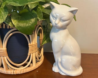 May include: A white ceramic cat figurine in a seated position with its tail curled around its side. The cat has pointed ears, almond-shaped eyes, and a smooth, glossy finish. The figurine is depicted in a static, upright posture.