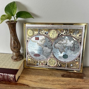 May include: A framed antique world map with a gold-colored frame. The map features detailed illustrations of continents and oceans in shades of blue, beige, and gold. A small vase with green leaves sits to the left of the frame.