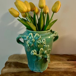 May include: A teal ceramic vase with embossed floral designs and handles, holding a bouquet of yellow tulips. The vase has a glossy finish and sits on a wooden surface, creating a vibrant contrast.