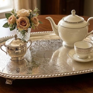May include: An oval silver serving tray with ornate embossed floral patterns and rope-style edges, featuring a white porcelain teapot, a matching teacup on a saucer, a silver sugar bowl with a lid, and a small glass vase of peach roses.