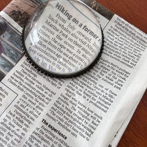 May include: A clear magnifying glass with a black metal rim is centered over a newspaper article. The article's headline reads "Hiking on a former". The newspaper is on a wooden surface.
