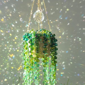 May include: A hanging plant ornament crafted from faceted green and yellow crystal beads, arranged to resemble a trailing vine in a small woven basket. The piece is suspended by gold-toned chains featuring a clear crystal sphere at the top.