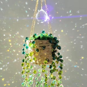 Può includere: Un cesto sospeso realizzato in materiale intrecciato di colore chiaro, riempito con muschio verde e fili a cascata di perline di cristallo sfaccettate nei toni del verde e del lime. Il cesto è appeso a catene dorate con un prisma di cristallo.
