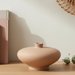 Może przedstawiać: Brzoskwiniowy ceramiczny wazon o zaokrąglonym korpusie i wąskiej szyi. Wazon stoi na drewnianej powierzchni, po lewej stronie książki i betonowy blok. W tle tkane lustro i zieleń.