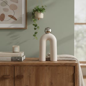 Peut inclure: Un objet décoratif en forme d'arche de couleur crème avec une sphère argentée sur le dessus. L'objet est exposé sur une commode en bois. Une petite bougie, des livres et une plante suspendue sont également visibles.