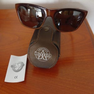 May include: Brown sunglasses with a dark brown frame and lenses, resting on a matching brown leather case. The case has the brand name "Vuarnet Sunglasses" printed on it. A white card with the brand name and "CE" is also present.