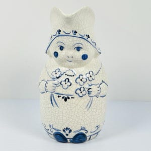 May include: A ceramic pitcher in the shape of a Dutch girl, featuring a white crackle glaze and blue accents. The pitcher has a spout, a hat, and holds a bouquet of flowers. The design is hand-painted with intricate details.