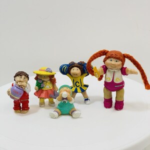 May include: A collection of vintage Cabbage Patch Kids figurines. The figures are dressed in colorful outfits, including dresses, overalls, and a cheerleader uniform. One figure holds a cookie jar. The dolls have painted faces and yarn hair.