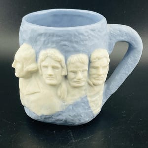 May include: A light blue ceramic mug featuring the sculpted faces of four U.S. presidents. The faces are in a cream color, contrasting with the blue mug. The mug has a handle and a textured surface.