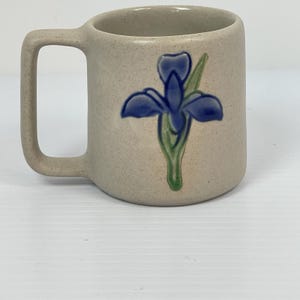 May include: A beige ceramic mug with a handle, featuring a blue iris flower design with green leaves. The mug has a speckled texture and a simple, rounded shape, suitable for hot beverages.