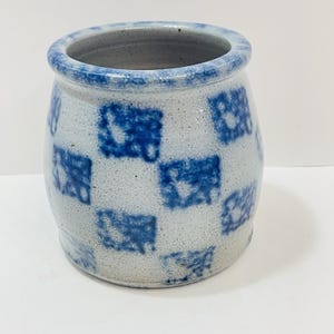 May include: A small, cylindrical stoneware jar with a light gray base and a repeating pattern of blue squares. The rim and the interior are also blue. The jar has a rustic, handmade appearance.