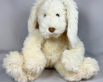 May include: A plush stuffed animal in the shape of a rabbit with long, floppy ears. The toy is covered in cream-colored, shaggy faux fur and features a small, brown, embroidered nose and dark, round eyes set into the soft, textured material.