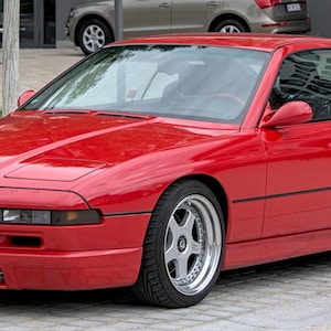 Puede incluir: Un cupé BMW Serie 8 rojo brillante con llantas de aleación plateadas. El coche tiene un diseño elegante y aerodinámico. La parte delantera presenta una parrilla negra y faros. La matrícula dice "BB NM 211".