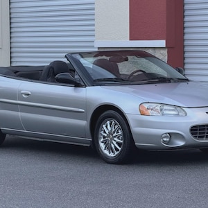 May include: Silver convertible car with a black interior and chrome wheels. The car is parked in front of a building with a gray garage door. The top is down, revealing the interior.