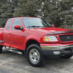Può includere: Un pick-up Ford F-150 rosso con cofano nero e dettagli cromati. Il camion ha una cabina equipaggio ed è parcheggiato su una superficie pavimentata. Il veicolo è di fronte a uno sfondo di alberi verdi.