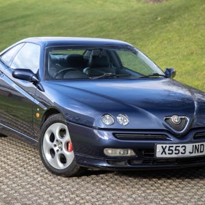Könnte beinhalten: Dunkelblaues Alfa Romeo Coupé, geparkt auf einer gepflasterten Oberfläche mit grünem Gras im Hintergrund. Das Auto hat silberne Räder, eine schwarze Innenausstattung und ein Nummernschild mit der Aufschrift "X553 JND".