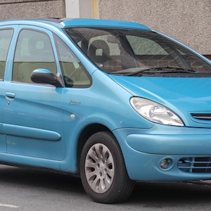 May include: A light blue Citroen Xsara Picasso. The car has a rounded body, with a large windscreen and side windows. The wheels are silver, and the car has a small spoiler above the windscreen. The word "Picasso" is visible on the side.
