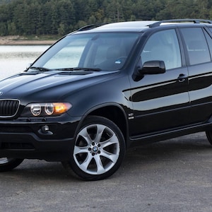 May include: A black BMW X5 SUV parked on a paved surface near a body of water. The vehicle has silver alloy wheels, a black grille, and tinted windows. The headlights are on, and the car is in good condition.