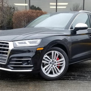 May include: A black Audi SUV with silver rims and red brake calipers. The vehicle is parked on a wet, paved surface. The front of the car features a chrome grill and headlights.