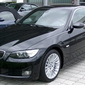 Op de afbeelding: Een zwarte BMW coupé met zilveren velgen staat geparkeerd op een grijs bakstenen oppervlak. De auto heeft een glanzende afwerking en een strak design. Een andere donkerblauwe cabriolet is gedeeltelijk zichtbaar op de achtergrond.