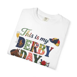 以下が含まれることがあります： 「This is my DERBY DAY」の文字がカラフルな模様で描かれた白いTシャツ。シャンパングラス、馬、ジョッキーの帽子、バラのデザインが施されており、競馬をテーマにしています。