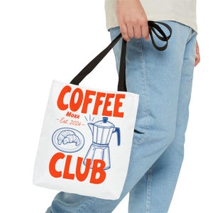 Puede incluir: Bolso tote blanco con asas negras. Muestra las palabras "COFFEE CLUB" en naranja, una ilustración de cafetera y croissant, y el texto "MOKA - Est. 2024".