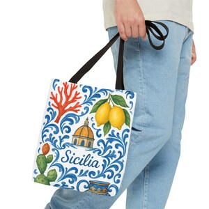 May include: A white tote bag with black straps featuring a blue scrollwork pattern, two yellow lemons, a red coral branch, a green prickly pear cactus, a small architectural dome, and a blue ceramic vessel with the word Sicilia printed in blue.