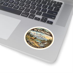 Op de afbeelding: Een ronde sticker met een landschapsillustratie van Wyoming. Het ontwerp toont bergen, een meer, een waterval en een hert. De tekst "WYOMING" en "THE EQUALITY STATE" staan op de sticker.