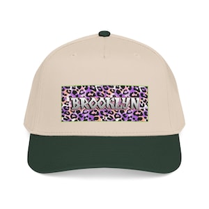 May include: A beige baseball cap with a dark green brim and a top button. The front features a rectangular patch with a purple, pink, and black leopard print background and the word BROOKLYN written in bold, metallic-style silver lettering.